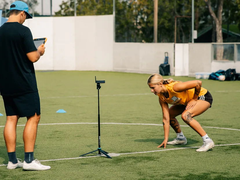 A young female athlete in a sprint-ready stance at a timing gate while a coach records her result